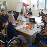 Die TeilnehmerInnen der Sitzung im Mensch Zuerst Büro in Götzis. Vor den SprecherInnen und SchreiberInnen: gefüllte Wassergläser und leere Kaffeetassen, vollgeschriebene Dokumente und Ordner, ein Laptop in der Mitte des Tisches. Im Hintergrund Ausblick au
