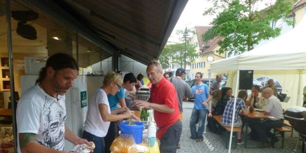 Die Essen und Getränkeausgabe beim Sommerreizfest. Im Vordergrund der Grill gefüllt mit bruzzelnden Köstlichkeiten. Die Gäste sitzen im Zelt, die Musik spielt auf, die Hungrigen und Durstigen sind bei der Ausgabe.