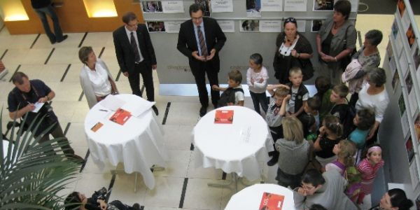 Eine Ausstellungseröffnung von oben betrachtet. Ein Redner formt die Hände zur Schale. Viele Köpfe, die zuschauen und zuhören. Ein Mädchen schaut herauf.