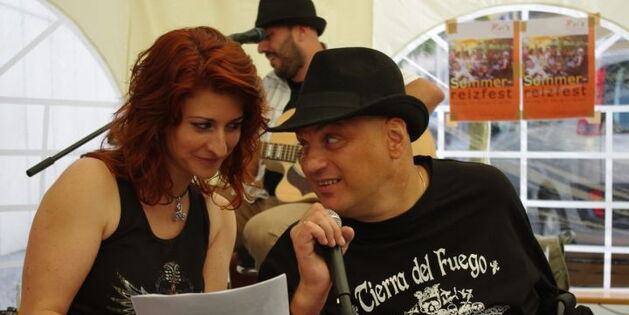 Die Leadsängerin von C.C.Mate Claudia singt mit Reinhard die Krüppellieder. Reinhard hält das Mikrofon, Claudia den Text. Beide schauen sich in die Augen. Im Hintergrund der Gitarrist und das Plakat zum Sommerreizfest.