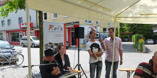 Obmann Andreas Guth bei der Eröffnungsrede. Reinhard Zischg übersetzt. Marianne Schulze lacht fröhlich. Auf dem Kopfsteinpflaster reflektiert die Sonne.