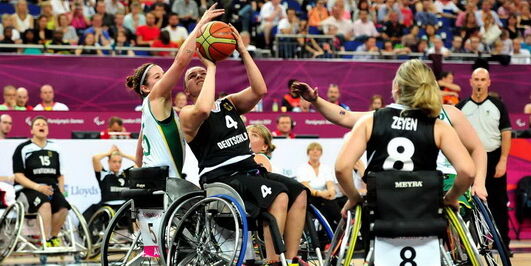 Basketballerinnen in Rollstühlen beim Spiel. Eine Frau versucht den Ball Richtung Korb zu werfen. Die Gegenspielerin im weißen Dress hält dagegen. Im Hintergrund Teammates und Zuschauerränge. Foto: backview.eu, Andreas Joneck