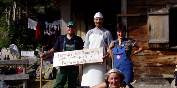 Alppersonal. Im Vordergrund kniet Sennerin Simone König bei der Ziege Brunhilde und umarmt das Tier. Dahinter stehen der Beisenn in weißer Schürze, ein Knecht mit Mistgabel und eine Magd mit Hütestecken. Alle lachen zum Betrachter.