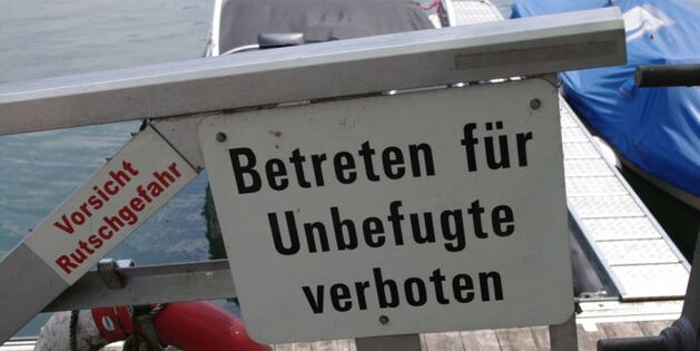 Auf einem Schild steht "Betreten für Unbefugte verboten". Und auf einem Geländer "Vorsicht Rutschgefahr". Im Hintergrund Wasser, Boote, ein Steg und Rettungsreifen.