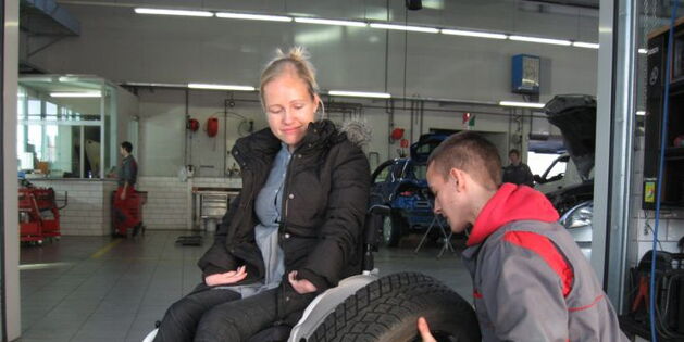 Reifenwechsel: Eine junge Frau im Rollstuhl schaut dem Automechaniker zu, der versucht einen Autoreifen auf ihrem Rollstuhl zu montieren.