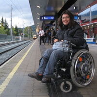 Die lachende Claudia Fessler in ihrem Elektrorollstuhl wartet auf dem Bahnsteig in Dornbirn auf den einfahrenden Zug.