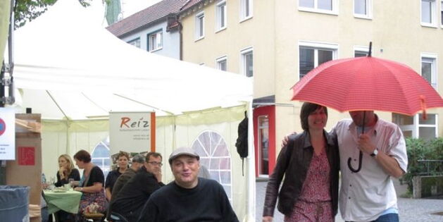 Ein Paar tanzt rot beschirmt durch den Regen. Der Ex rollt lachend dazu. Im Hintergrund schauen die Festgäste aus dem Zelt.
