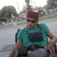 Ein Rollstuhlfahrer als Tourist in Istanbul. Mit Sonnenbrille vor den Augen und weinrotem Fez auf dem Kopf. Im Hintergrund zwischen Palmen und Bäumen die vier Türme der blauen Moschee.
