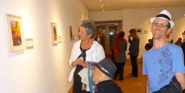 Eine Frau und ein Rollstuhlfahrer mit schwarzem Hut am Kopf betrachten das Bild "Schale mit Kristallstein" von Elfi Plankensteiner. Ein Mann mit weißem Hut am Kopf lächelt in die Kamera. Im Hintergrund diskutierende Ausstellungsbesucher.