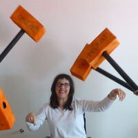 Eine Rollstuhlfahrerin. Mehrere orangefarbene Hämmer sausen auf sie. Sie lässt sich nicht beeindrucken und lacht in die Kamera.