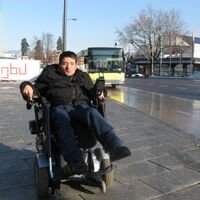 Sueleyman Kurt wartet in seinem Elektrorollstuhl auf den einfahrenden gelben Landbus. Im Hintergrund blauer Himmel und das Kulturhaus Dornbirn.