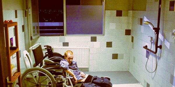 Ein Badezimmer. Ein Mann ist ausgerutscht und kauert am Boden. Mit einer Hand stützt er sich am Rollstuhl und wartet auf Hilfe.