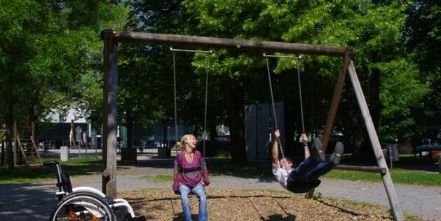 Ein Mann schaukelt mit Beinen voraus hoch hinaus. Eine Frau auf der Nebenschaukel lacht mit Kopf im Nacken hoch hinaus. Ein weißgerahmter Rollstuhl und saftiggrüne Parkbäume sehen zu.