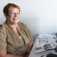 Eine freundlich lachende Frau sitzt vor ihrem Büroschreibtisch und schaut in die Kamera. Rechner, Tastatur, Maus, Schreibstifte und ein Bildschirm schmücken den Tisch. Auf ihren Arbeitspapieren liegt die Brille.