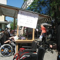 Ein Plakat an einem Marktstand kündigt Rollstuhlselbsterfahrung an. Mehrere RollstuhlfahrerInnen sind am Stand beschäftigt. Leere Rollstühle stehen bereit. Ganz rechts ein Mann mit Sonnenbrille lädt ein zum Sitzen.