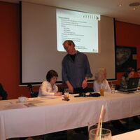 Am Podium links sitzen Reinhard und Julia, die das Protokoll führt. Eberhard steht als Moderator in der Mitte und spricht zum Publikum. Links von ihm sitzen Angi und Andi und lauschen Eberhards Vortrag.