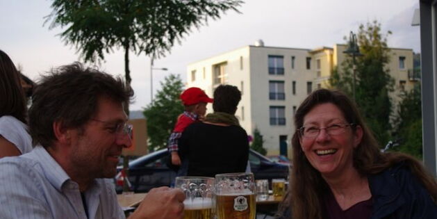 Ein Mann und eine Frau stoßen mit ihren Biergkrügen an. Die Frau schaut mit einem fröhlichen Lachen in die Kamera. Der Mann konzentriert sich auf die Bierkrüge.