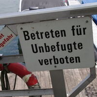 Auf einem Schild steht "Betreten für Unbefugte verboten". Und auf einem Geländer "Vorsicht Rutschgefahr". Im Hintergrund Wasser, Boote, ein Steg und Rettungsreifen.