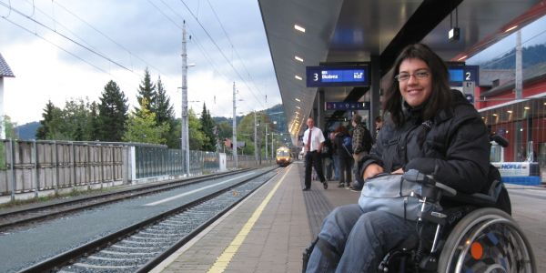 Die lachende Claudia Fessler in ihrem Elektrorollstuhl wartet auf dem Bahnsteig in Dornbirn auf den einfahrenden Zug.