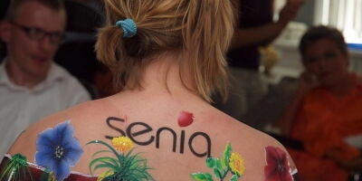 Foto: Verein Senia. Eine Frau mit dem Rücken zum Betrachter. Blonde Haare mit einem blauen Band zusammengebunden. Der Rücken bis zur Schulter bemalt mit Ziegelsteinen und bunten Blumen.