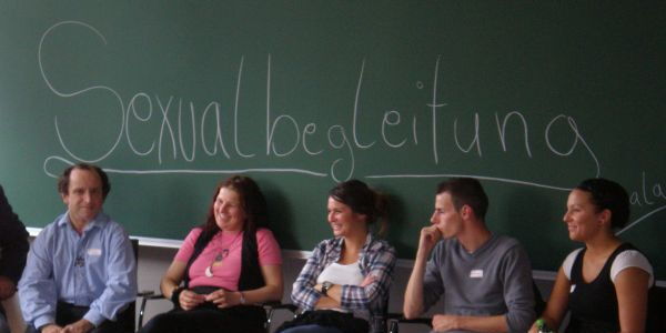 An einer großen Tafel steht mit weißer Kreide das Wort Sexualbegleitung. Vor dieser Tafel sitzt die Workshopleiterin Vimala mit mehreren Teilnehmern - manche lachend, manche schmunzelnd.