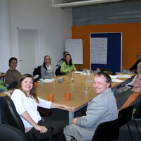 Das Bild zeigt den Büroraum in Bludenz mit der Reiz-orangen Wand im Hintergrund. Die Teams von Mensch Zuerst-People First Vorarlberg und Reiz sitzen um einen großen Tisch. Alle schauen fröhlich in die Kamera.