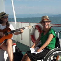 Auf dem Schiff am Bodenssee. An der Reling sitzen zwei Frauen und singen. Gitarre und Liederbuch in der Hand. Gelbleuchtende Strohhüte auf dem Kopf. Strahlendes Lachen. Im Hintergrund segeln weißblinkende Boote.