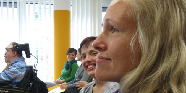 Im Vordergrund sieht man groß das Profil von Sabrina Nitz. Dahinter lächelnd schaut Julia Fischbach in die Kamera. Im Hintergrund ein Rollstuhlfahrer und mehrere KongressteilnehmerInnen beim Smalltalk.