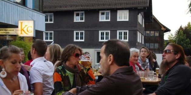 Viele Menschen sitzen entspannt oder fröhlich an Biertischen. Ein Mann und eine Frau im Vordergrund essen Grillspezialitäten. Eine Frau im Hintergrund trinkt aus dem Bierkrug.