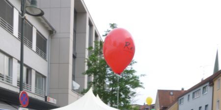 Julia Fischbach, junge hübsche Frau mit rötlichen Haaren, schaut fröhlich in die Kamera. Sie hält einen rot leuchtenden Luftballon mit Postkarte startbereit in die Höhe.