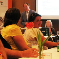 Im Hintergrund links der Reiz-Banner. Eberhard Zumtobel moderiert die Generalversammlung. Sabrina Nitz und Julia Fischbach sitzen neben ihm auf dem Podium des Reiz-Vorstands. Im Vordergrund Publikum am Tisch und eine rotblühende Blume in einer Vase.