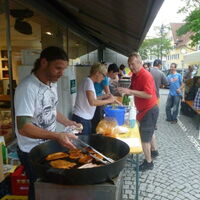 Die Essen und Getränkeausgabe beim Sommerreizfest. Im Vordergrund der Grill gefüllt mit bruzzelnden Köstlichkeiten. Die Gäste sitzen im Zelt, die Musik spielt auf, die Hungrigen und Durstigen sind bei der Ausgabe.