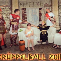 Der Krüppelballflyer 2016. Ein römischer Soldat kitzelt mit seiner Lanze den ägyptischen Pharao. Göttin in Weiß sitzt verträumt auf der Couch. Cäsar mit Amphore in der Hand und Jesus im Rollstuhl schauen zu. Die römische Soldatin schaut grimmig.