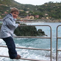 Ein Junge mit kritischem Blick hält sich am Geländer fest. Unter ihm Wellengischt. Im Hintergrund ein Felsen im Wasser, Sandstrand und ein Dorf am ligurischen Meer.