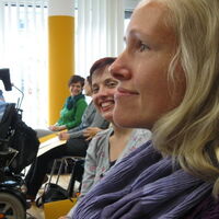 Im Vordergrund sieht man groß das Profil von Sabrina Nitz. Dahinter lächelnd schaut Julia Fischbach in die Kamera. Im Hintergrund ein Rollstuhlfahrer und mehrere KongressteilnehmerInnen beim Smalltalk.