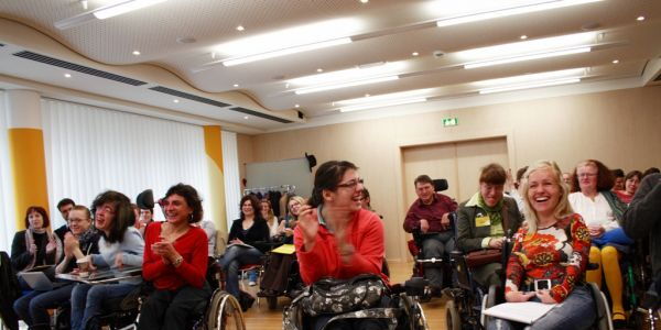 Foto: Bizeps. Gute Stimmung bei den KongressteilnehmerInnen. Viele lachende Gesichter und applaudierende Hände. Im Vordergrund Claudia Fessler und Sabrina Nitz in ihren Rollstühlen.