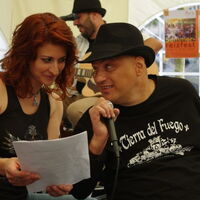 Die Leadsängerin von C.C.Mate Claudia singt mit Reinhard die Krüppellieder. Reinhard hält das Mikrofon, Claudia den Text. Beide schauen sich in die Augen. Im Hintergrund der Gitarrist und das Plakat zum Sommerreizfest.