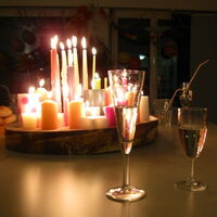Auf einer großen Holzscheibe leuchten viele brennenden Kerze: große und kleine, weiße und bunte. Ein paar Sektgläser stehen am Tisch um das Kerzenlicht herum. Aus dem Schatten am rechten Bildrand lächelt eine fröhliche Frau.