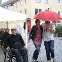 Ein Paar tanzt rot beschirmt durch den Regen. Der Ex rollt lachend dazu. Im Hintergrund schauen die Festgäste aus dem Zelt.