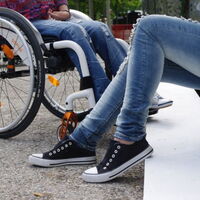 Zwei Beinpaare im Gespräch? Ein Beinpaaar in Jeans mit blauweißen Turnschuhen. Angewinkelt in Sitzposition auf weißen Stufen. Ihm zugewandt das andere Beinpaar. Blaue Jeans, hellgraue Turnschuhe, im Rollstuhl mit weißem Rahmen und schwarzen Reifen.