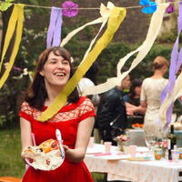 Eine junge Frau in rotem Kleid trägt mit glüchlichem Lachen ihren Teller voll Kuchen zwischen bunten Girlanden. Im Hintergrund gedeckte Tische und Partygäste einer Gartenfeier. Fotoquelle: Alamode