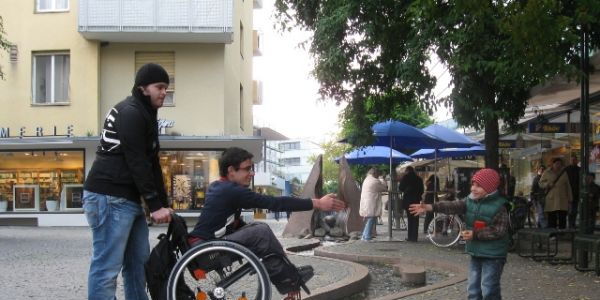 Ein Rollstuhlfahrer steht mit Hilfe eines Assistenten auf einem kleinen Steg über einem Wasserlauf. Er streckt die Hand aus. Auf der anderen Seite des Wasserlaufs steht ein Kind, das die Hand nach ihm streckt. Komm!