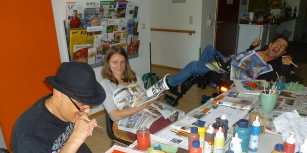 Ein Tisch bunt befüllt mit Malfarbentuben. Zeitungspapier quillt unter Kunstwerken hervor. Reinhard mit schwarzem Hut setzt den Malstift an. Katrin schenkt dem Kameramann ein Lächeln, Sülo verschenkt ein Lachen.