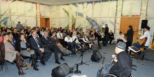 Foto: VLK/A. Serra. Rechts im Vordergrund singt Reinhard Zischg das Siegerlied, begleitet von Matthias Zabransky an der Gitarre. Im Hintergrund das Publikum bei der Chancenpreisverleihung 2011. Ein vollbesetzter Montfortsaal.