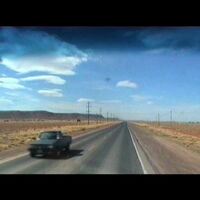 Blick aus dem Auto: Blauer Himmel mit spärlichen Wolken. Sand, kaum Pflanzen. Eine lange gerade Straße verschwindet am Horizont. Ein Pickup kommt entgegen, sonst ist die Straße frei.