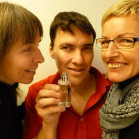 Ein rotbehemdeter Mann hält die Parfumflasche des Duftes Reiz Unisex in der Hand. Zwei Frauen schnuppern daran.