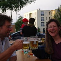 Ein Mann und eine Frau stoßen mit ihren Biergkrügen an. Die Frau schaut mit einem fröhlichen Lachen in die Kamera. Der Mann konzentriert sich auf die Bierkrüge.