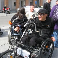 Ein Rollstuhlfahrer in seinem Bike. Schwarzes Outfit und Fahrradbrille. Blick in Aufbruchstimmung und Fahrtrichtung, eine Hand am Tretlenker, bereit zur Abfahrt.