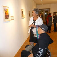 Eine Frau und ein Rollstuhlfahrer mit schwarzem Hut am Kopf betrachten das Bild "Schale mit Kristallstein" von Elfi Plankensteiner. Ein Mann mit weißem Hut am Kopf lächelt in die Kamera. Im Hintergrund diskutierende Ausstellungsbesucher.