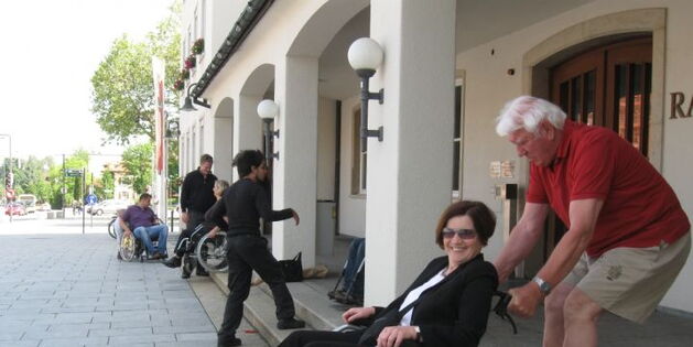 Ein Mann mit weißen Haaren zieht konzentriert eine Rollstuhlfahrerin samt Stuhl über Treppenstufen hoch. Die Rollstuhlfahrerin lacht gespannt in die Kamera.